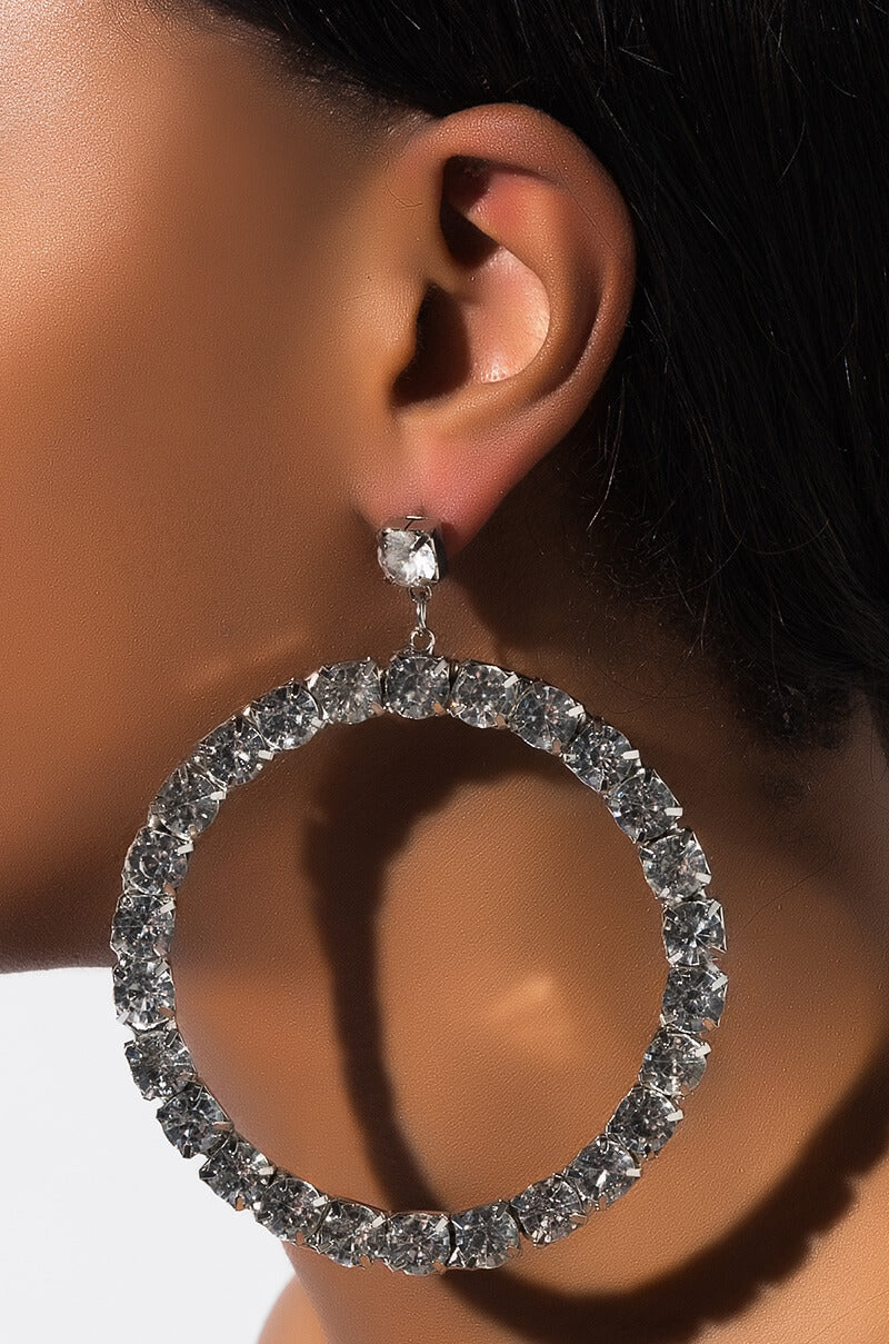 Close-up of a person's ear wearing the TRY ME RHINESTONE HOOP EARRING by Y&S Davidson Inc, a large, round hoop earring adorned with sparkling rhinestones. The light reflects off the stones, creating a shimmering effect. The person's skin tone is warm, and the background is a light, neutral color.