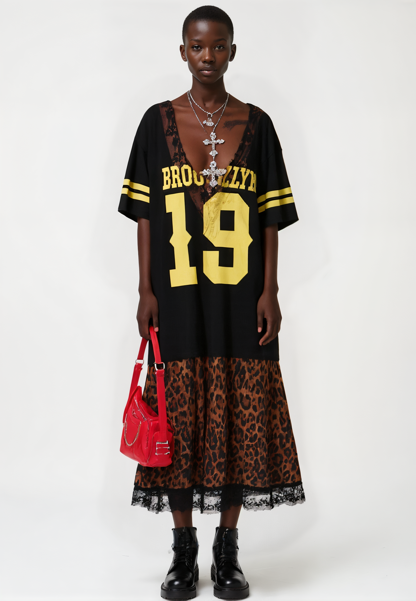 A person in the CITY GIRL LAYERED JERSEY MAXI DRESS with "BROOKLYN 19" in yellow, leopard print hem, black boots, layered necklaces, and a shiny red handbag stands against a white background.
