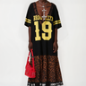 A person poses against a white background in the CITY GIRL LAYERED JERSEY MIDI DRESS—black mesh with "BROOKLYN 19" in yellow, leopard print hem, black lace trim—paired with chunky boots, layered necklaces, and a red handbag.