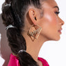 A woman with a long braided ponytail tied with silver bands is shown in profile. She is wearing a pink, silky material and the VIRGO PAVE HOOP earrings, which are large gold square hoops that spell "Unique." Her makeup includes a subtle highlighter and soft pink lipstick.