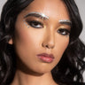 A person with wavy dark hair and shimmering makeup. They have rhinestones from the VIXEN EYE GEM LINER above the eyebrows and below the eyes, and they wear glossy nude lipstick. Their expression is neutral, and light catches on the rhinestones from the VIXEN EYE GEM LINER above their right eyebrow, creating a starburst effect.