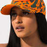 A person with long, dark hair is wearing a VON DUTCH TRUCKER HAT ORANGE CAMO. They have a neutral expression and are looking slightly upwards, with a plain white background behind them.