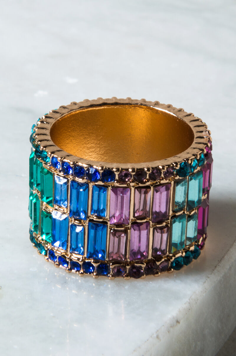 The WEDDING BARBIE COCKTAIL RING ORANGE GOLD is a stunning gold ring embellished with rows of rectangular gemstones in an array of colors such as blue, green, pink, and purple. These vibrant stones are meticulously set in vertical columns around the band, giving the ring a dynamic and sophisticated look against a light marble surface background.