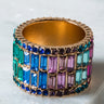 The WEDDING BARBIE COCKTAIL RING PINK MULTI is a wide, gold-toned ring encrusted with rectangular gemstones meticulously arranged in rows. The vibrant stones come in various colors including blue, green, pink, and purple, forming a striking mosaic-like pattern against its smooth, light-colored background.