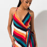 A woman with long black hair tied in a high ponytail is wearing the SEXY MULTI COLOR STRIPE DRESSY MINI, which features a sleeveless, V-neck wrap design with vibrant diagonal stripes. She has her hands by her sides and is looking slightly downward against a plain white background.