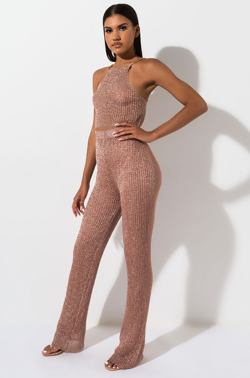 A woman stands against a white background wearing the WEIGHT IN GOLD PANT, a sleeveless, ribbed knit rose gold jumpsuit. The outfit features a high neckline, fitted bodice, and wide-leg pants. She poses with her hand on her hip, showcasing the full length of the sparkling ensemble.