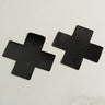 Two DOM-XF-NS adhesive patches, each shaped like a black cross, are arranged side by side on a light gray surface. The patches boast a matte finish with sharp, clean edges and feature a smooth and even surface texture.