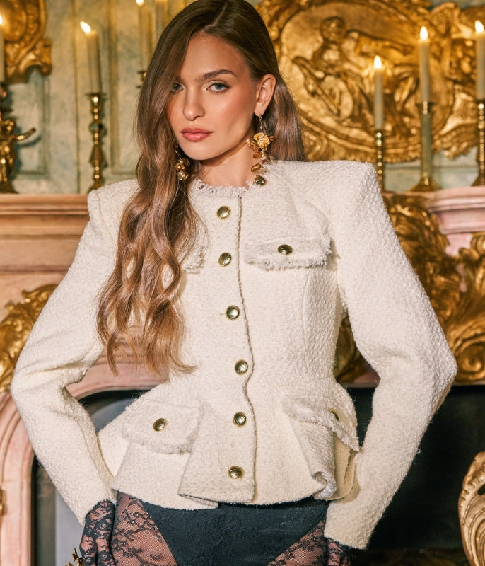 A woman models a cream-colored tweed jacket with gold buttons and flap pockets, set against an ornate, candle-lit gold interior.
