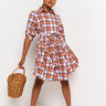 A person stands confidently, wearing the YOU NEED BUTTON DOWN MINI DRESS, featuring a striking colorful plaid pattern with a tied waist. They hold a wicker handbag in one hand and wear clear high heels. The background is plain white.